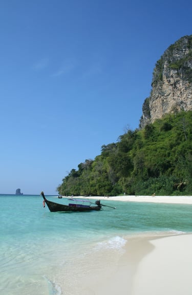 A traditional Thai longtail boat floats in turquoise water by a white sand tropical beach and limestone cliffs.