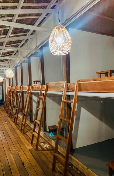 Rustic wooden capsule dorm beds with ladders and wicker pendant lighting in a cozy tropical hostel.