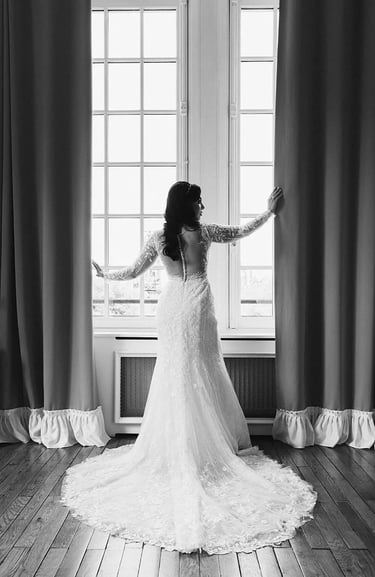Mariée se tenant près d'une fenêtre et portant une magnifique longue robe blanche.