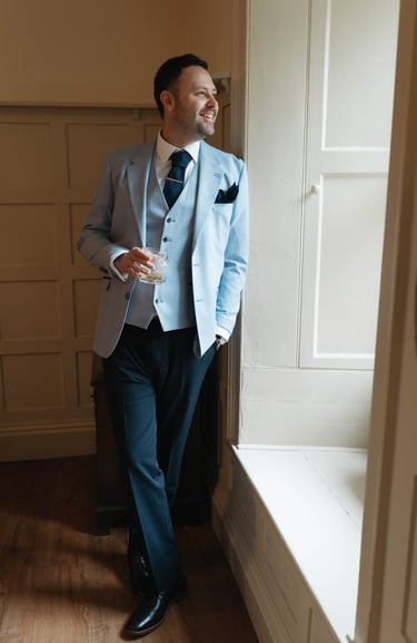 Groom in light blue three-piece windowpane suit holding whiskey by a window in glasgow.