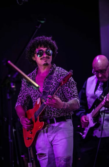 A guitarist with curly hair and sunglasses performs live music on a red electric guitar at a concert venue.