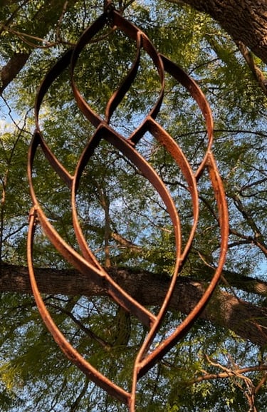 Rustic metal leaf garden sculpture hanging from a green tree canopy outdoors.