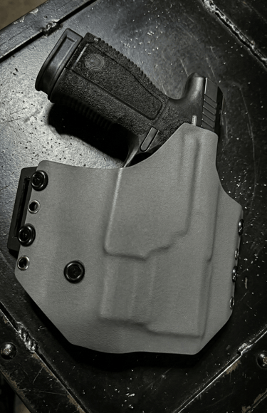 A black semi-automatic handgun secured in a custom gray Kydex OWB holster on a textured surface.