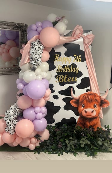 Festive birthday decorations with a cowhide pattern, pink and purple balloons, lush greenery, and a cutout of a cute