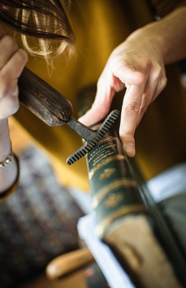 Artisan using a vintage bookbinding wheel tool to emboss gold details onto a leather book spine.