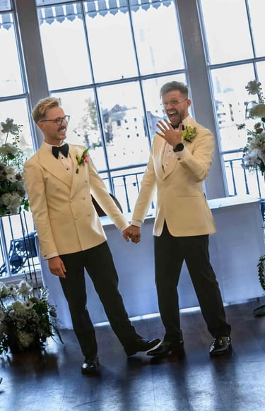 Two grooms in cream tuxedos holding hands and celebrating after their indoor wedding ceremony.