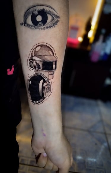 Tatuaje de los cascos de Daft Punk en un antebrazo, debajo de un tatuaje de un ojo surrealista.