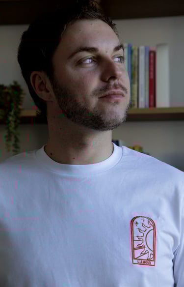 Young man with a beard wearing a white t-shirt featuring a red embroidered L'Amour sun graphic.