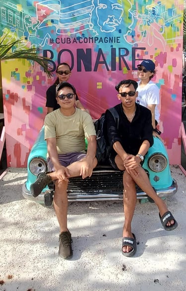 Four people posing on a vintage turquoise car in front of a colorful Cuba Compagnie Bonaire mural.