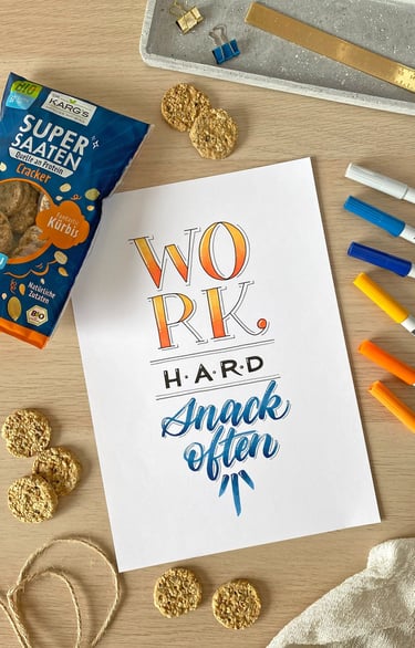 Hand-lettered motivational quote on a desk with colorful markers and healthy pumpkin seed crackers.