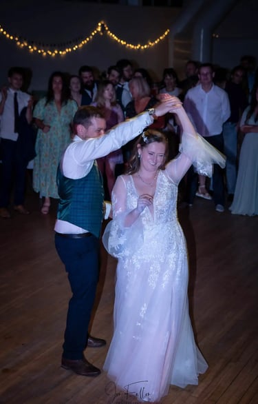 A smiling bride in a lace gown and groom in a green vest dance their first dance at a wedding reception.