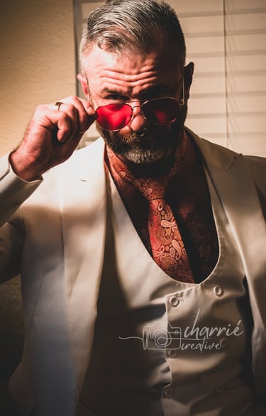 dapper bearded man dressed in formal clothing in low light staring intently over his glasses
