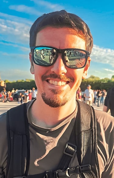 Portrait d’un homme souriant avec des lunettes de soleil reflétant une foule, ciel bleu et ambiance ensoleillée d’événement