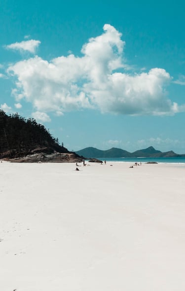 Pristine white sand beach with tourists, turquoise ocean water, and lush tropical hills under a blue sky.