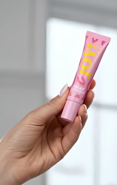 A hand holding a pink tube of Joy beauty lip gloss or skin cream against a blurred background.