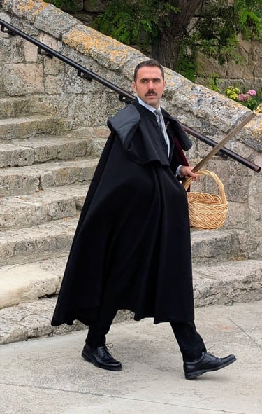 Cofradía member in black cape and suit with red tie, holding a basket and a sword, standing on stone steps during El Colacho
