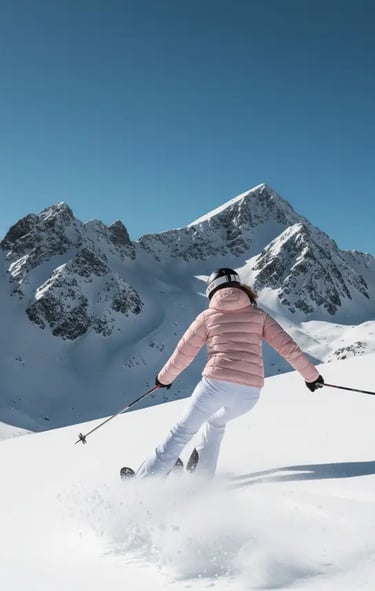 Female skier in a soft pink jacket and white trousers skiing down a mountain slope under a clear blu