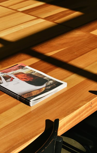 A stack of magazines on a light wood table with natural sunlight and window shadows.
