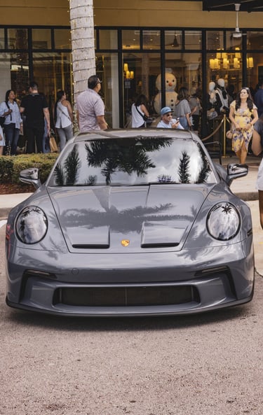 Front view of a grey Porsche 911 GT3 parked outside a high-end shopping center with palm tree reflections.
