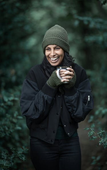 Shivani wearing winter clothes, grinning, holding a mug and standing in woodland.