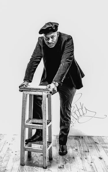 Portrait en noir et blanc de Teddy Bob, photographe lifestyle, posant avec assurance derrière un tabouret. Vêtu élégamment