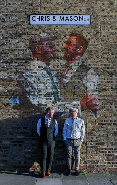 Gay couple posing under a custom street sign and mural of themselves on a London brick wall.