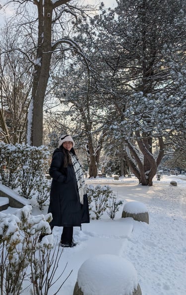 A woman in a long black winter coat and scarf stands in a snow-covered park surrounded by frosted pine trees.