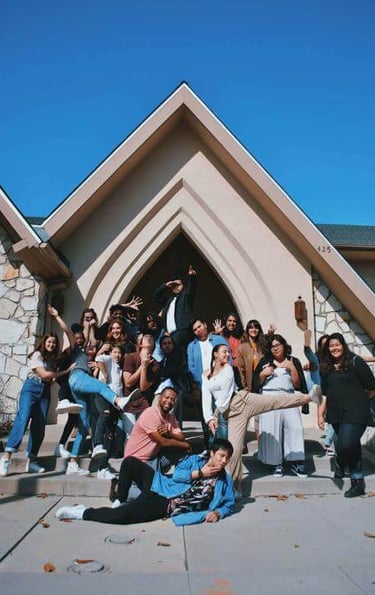 Energetic group striking playful poses in front of a church-like building under a bright blue sky.