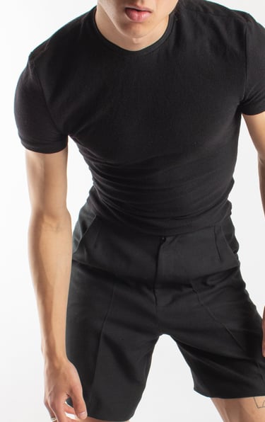 A man wearing a slim-fit black t-shirt and pleated trousers in a studio setting.