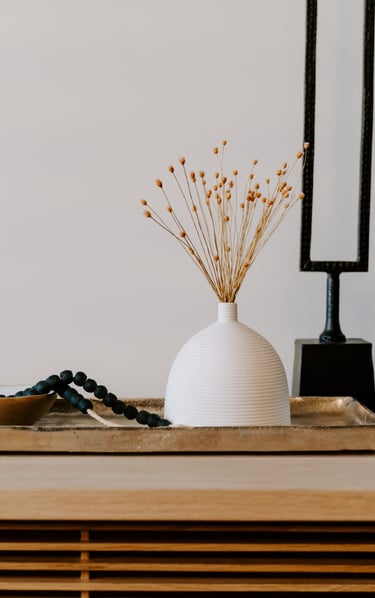 Minimalist home decor featuring a white ribbed vase with dried flowers on a gold tray with black beads.