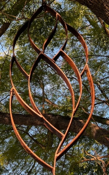 Rustic metal leaf garden sculpture hanging from a green tree canopy outdoors.