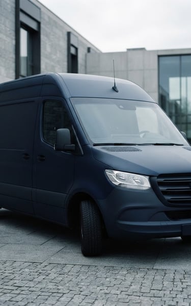 A pristine, unmarked dark transport vehicle parked outside a modern stone facility under soft overcast daylight, cool silver tones, shallow depth of field.