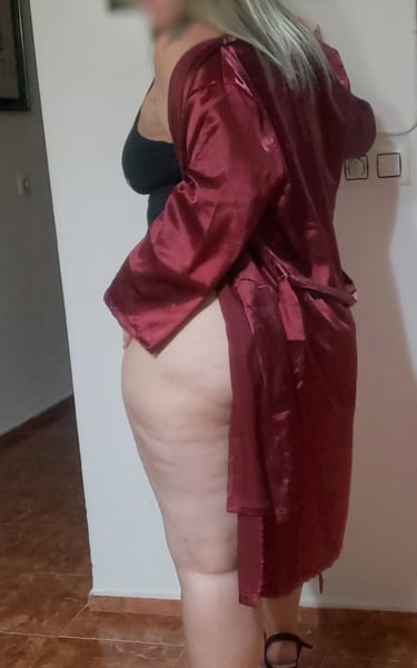 Escort madura colombiana en bata roja de satén, de rodillas sobre el sofá, mostrando sus piernas provocativas.