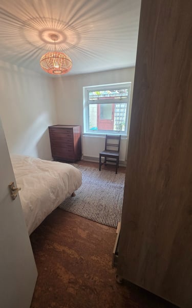 A bright bedroom with a woven pendant light, wooden dresser, and window view of a patio.