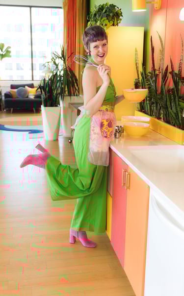 A woman in a green jumpsuit and pink boots baking in a colorful mid-century modern kitchen.