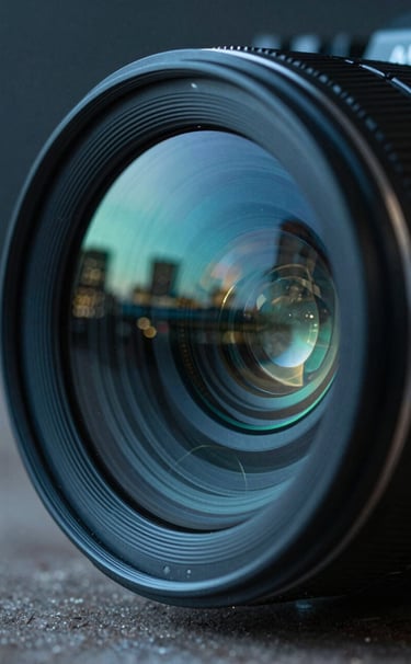 A sharp, cinematic close-up of a high-end film camera lens. The glass reflects a blurred metropolitan skyline at blue hour, featuring tones of deep midnight teal and muted seafoam green.
