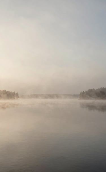 A serene wide-angle landscape photograph of a misty lake in Ontario, Canada, at dawn. The water is perfectly still, reflecting a soft beige and muted grey sky. Minimalist photography style with ample negative space.