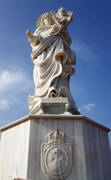 White marble statue of Virgin Mary holding Child Jesus against a blue sky background.