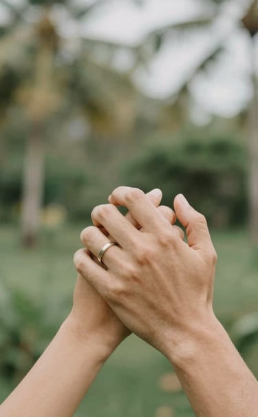 Close-up of a couple's hands intertwined, wearing simple rings, with a beautifully blurred tropical background in Bali. Warm, intimate storytelling aesthetic, natural lighting, featuring tones of #5F705B, #C7B7A3, and #F7F3EE.