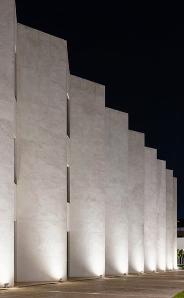 A wide, cinematic shot of a modern architectural facade in a South American / Brazilian setting. The building is composed of white geometric blocks against a pure black night sky. Elegant exterior lighting highlights the texture of the materials.