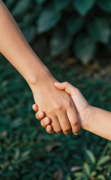 Candid close-up of a child's hand holding an adult's hand, softly lit by warm sunlight. The background is a blurred garden with deep teal green foliage and a few charcoal shadows for depth.