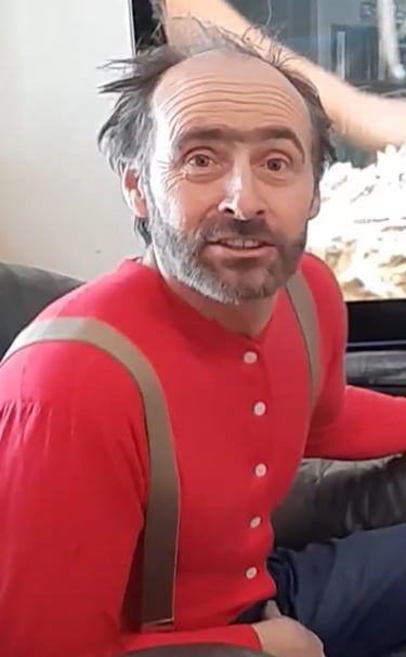 Hank with messy hair and a beard wearing a red union suit with suspenders indoors.