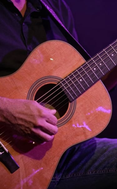 Close-up of a musician's hands playing an acoustic guitar with intensity, shot in a dimly lit South American venue with deep purple and rose colored ambient lighting reflecting on the wood.