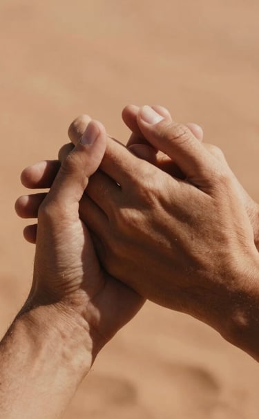 A warm, close-up photograph of two hands gently holding each other. Soft, warm sunlight filtering through, sandy textured backdrop, rich terracotta tones, intimate cinematic feel, Spain / Southern Europe.