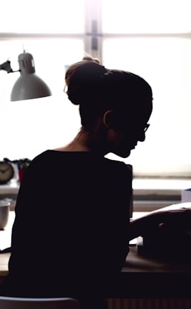 Rearview silhouette of a young woman sitting at a desk in front of a window