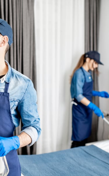 Personal de limpieza de hombre y una mujer con uniforme azul, limpiando habitación.