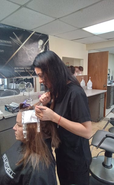 A student at a beauty school applying hair highlights with foil to a client in a salon setting.