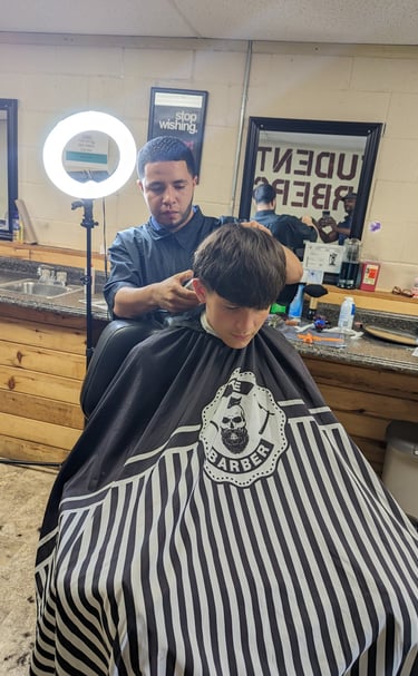 A student barber giving a haircut with electric clippers in a barber college setting.