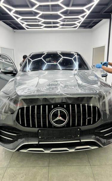 A technician applies clear paint protection film to a grey Mercedes-Benz SUV hood in a modern auto detailing shop.