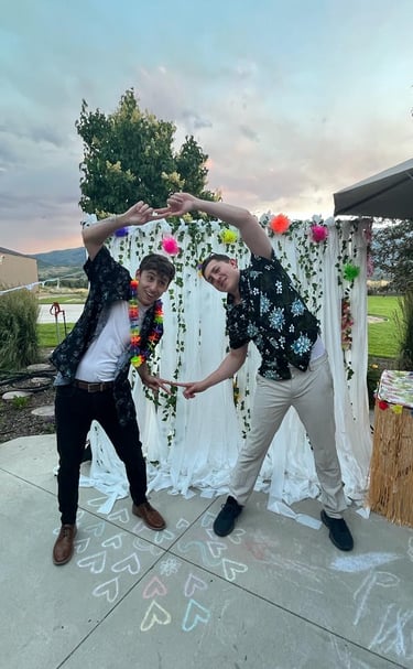Two men in Hawaiian shirts make a heart shape at a tropical theme outdoor party.